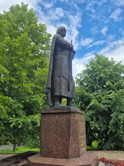 This media file captures a bronze statue of a person in historical armor, holding a sword. The statue stands on a large, multi-faceted granite pedestal with visible inscriptions. The background is dominated by lush green trees and a bright blue sky with scattered white clouds.

The statue appears to be a monument, possibly commemorating a historical figure. The person depicted is wearing a chainmail-like armor and a helmet, suggesting a warrior or a ruler from a past era. The sword is held upright, adding to the imposing presence of the statue.

The granite pedestal is rough in texture and has a speckled pattern of red and black. Faint inscriptions can be seen on one of its surfaces, although the text is not clearly legible in the image. The monument is situated outdoors, amidst a natural setting with abundant greenery.

The weather appears to be pleasant, with a clear blue sky and sunlight filtering through the leaves. The time of day is likely daytime, given the brightness of the sky. The overall scene evokes a sense of historical significance and natural beauty.

The location is identified as Petrozavodsk, Russia, which provides geographical context for the monument and its potential historical connections. The image is oriented vertically, giving a dramatic perspective of the statue against the sky and trees.