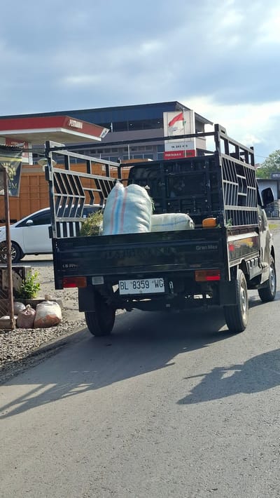 A black Daihatsu Gran Max pickup truck is parked on an asphalt road in the city of Keudebayu, Indonesia. The truck is loaded with two large sacks of what appears to be agricultural produce or fertilizer. The sacks are white with red and blue stripes. The truck has a metal cage bed. The license plate of the truck reads "BL 8359 WG A". In the background, a Pertamina gas station canopy is visible, with a red Indonesian flag flying above it. A white sedan is parked to the left of the truck. The road is shaded by the truck and objects on the left side of the frame. The sky is overcast.