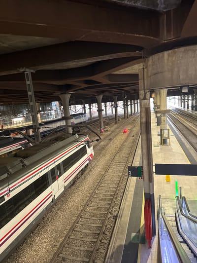 This image depicts the interior of the Oviedo/uviéu train station in Spain. The scene is dominated by train tracks and platforms, with a train visible on the left side of the frame. The train is white with red and purple stripes along its side. The station has a complex overhead structure of beams and columns, suggesting a large, multi-level facility. Several other trains and tracks are visible in the background. A sign with the number "3" and the word "Via" is positioned on the platform. Escalators are visible on the right side of the image, indicating access to different levels. The lighting suggests it is either early morning or late evening, or an indoor environment with artificial lighting. There are no people clearly visible in the image.