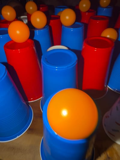 The image depicts a close-up, slightly blurred view of several orange ping pong balls resting on top of blue and red plastic cups. The cups are arranged in a seemingly haphazard manner on a wooden surface. There are no people visible in the image. The lighting is somewhat dim, casting shadows and highlighting the glossy surfaces of the balls and cups. The setting appears to be indoors, potentially a room or a table setup for a game. The image does not provide any specific information or context related to the city of Heijen, Netherlands.