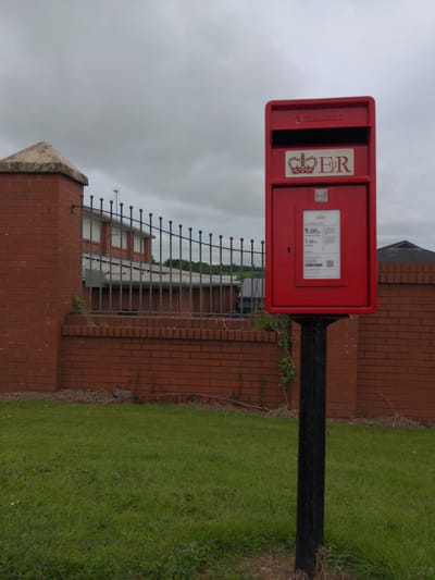 A red Royal Mail postbox stands on a black metal pole in front of a brick wall in Magherafelt, United Kingdom. The postbox has a Royal cypher and the letters "ER" on it. A sign on the postbox indicates the last collection times for mail. Beyond the postbox and wall is a patch of green grass and a building with a metal fence. The sky is overcast.