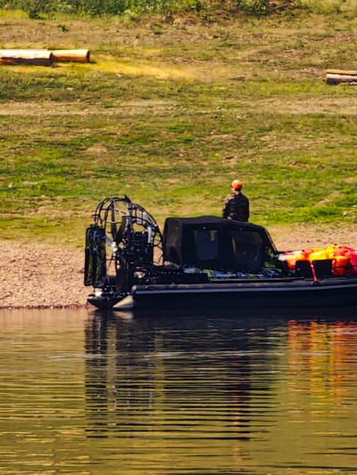 An airboat with a fan engine is docked on the shore of a body of water. A person wearing a camouflage jacket and an orange hat is standing on the back of the airboat. Several bright orange and yellow life jackets are stacked on the rear deck of the boat. The background consists of a grassy slope with some fallen logs. The water in the foreground is rippled, reflecting the boat and the surrounding environment. The image appears to be taken in Pogodaevo, Russia.