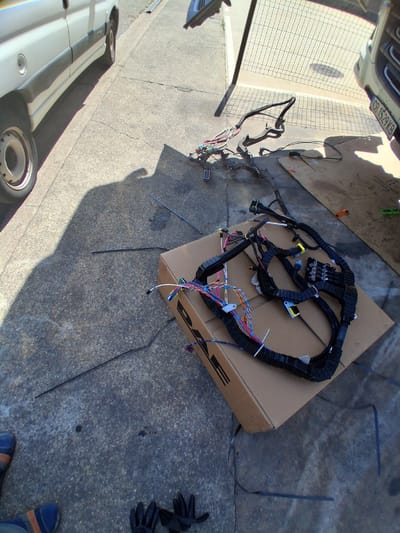 The image shows a car's wiring harness laid out on a cardboard box on a concrete surface. The scene appears to be outdoors, with sunlight casting shadows, suggesting it is daytime. The weather seems to be fair.

The main subject is the complex wiring harness, which is black and organized with various colored wires and connectors. It is partially spread out on a brown cardboard box that has a logo on its side. Some wires are neatly bundled, while others are more spread out, indicating it might have been recently removed from a vehicle or is being prepared for installation.

In the background, a portion of a white vehicle is visible on the left side of the frame, with its license plate reading "GB 1529 KB". The ground is a textured concrete surface with some dirt patches and cracks. To the right, a pair of black gloves and a shoe with blue and orange accents are visible, possibly belonging to the person working on the wiring harness.

The setting appears to be a driveway or an outdoor workshop area, possibly in Mas Aliu, Spain, given the location context. The overall impression is one of automotive repair or maintenance work being carried out outdoors. There are no clear indications of emotions, but the organized layout of the harness suggests a methodical approach to the task.