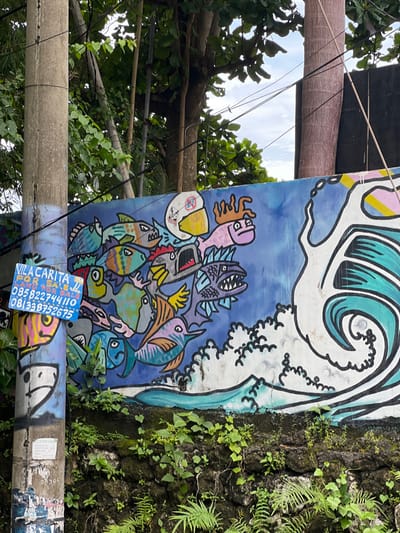 A vibrant mural displays stylized sea creatures in a dynamic composition against a blue background with a white wave at the top. Various fish, including sharks and swordfish, are clustered together. The mural is affixed to a textured wall covered in foliage. In the foreground, a gray pole with a blue business card advertising "Vila Carita" is visible. Wires and tree branches with green leaves frame the image on the left.