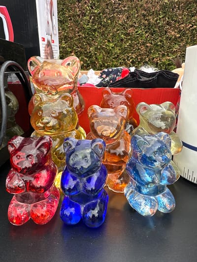 This image captures nine transparent, multi-colored figurines, each shaped like a gummy bear, displayed on a dark, reflective surface in Deinste, Germany. The figurines are arranged in two rows. The foreground features three distinct figures: one deep red, one dark blue, and one light blue. The back row contains six additional figurines, partially obscured by those in front, predominantly in amber, yellow, and orange tones, with one appearing light green-yellow.

Behind the figurines, a red cardboard box is visible, filled with assorted textiles, including a dark fabric with a floral pattern and a mass of white, fluffy material. To the immediate left of the red box, a segment of a black object with a red detail and a metallic, curved handle is present, along with a white box featuring an illustration of a white household appliance, possibly a blender or food processor. To the right of the red box, a white, ribbed cylindrical object stands. The background of the scene is dominated by a dense, green hedge, showing some areas of browning foliage, indicating an outdoor setting during daylight. No people are visible, and no actions or interactions are depicted.