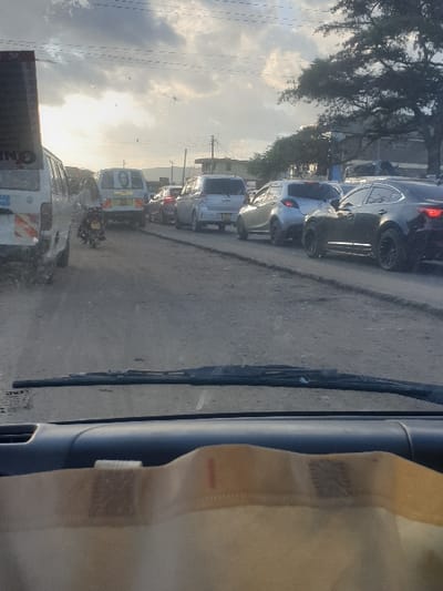 The scene depicts a busy, dusty road in Mumbuni North ward, Kenya, viewed from inside a vehicle. A significant traffic jam extends into the distance under a partly cloudy sky with visible sun towards the left.
The foreground shows the vehicle's dark dashboard and dirty windshield with wipers. A beige fabric object, possibly a bag, rests on the dashboard.
On the road, a line of diverse vehicles is stationary or moving slowly. Immediately to the left, a white van with blue accents and a red/white chevron marking is partially visible. Behind it, a motorcycle with a helmeted rider is positioned on the unpaved surface. Further down the lane, another white van, featuring a prominent portrait on its rear door, is visible. It is followed by a silver minivan/wagon, a lighter silver compact car, and several dark-colored sedans, all queuing in the traffic. Utility poles, overhead power lines, and sparse trees line the right side of the road, with indistinct buildings in the background. The road surface appears unpaved, uneven, and covered in dirt/dust.