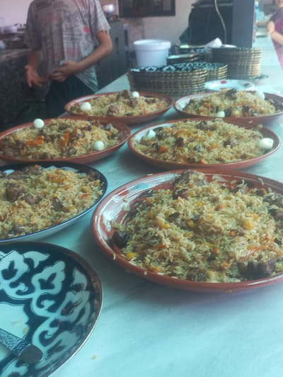 This is a medium-shot, eye-level photograph taken at eye-level inside what appears to be an outdoor restaurant or food stall. The scene is set in Yorkatay, Uzbekistan. A person, partially visible from the waist down, is standing in the background wearing a grey t-shirt and dark pants. The foreground is dominated by several oval-shaped serving dishes filled with pilaf, a traditional Uzbek rice dish. The pilaf contains rice, meat chunks, and shredded carrots, and some dishes are garnished with quail eggs. To the left, a patterned bowl with a blue and white design is visible, containing a fork. In the background, stacked plates and serving baskets are arranged on a table. The overall impression is one of a prepared meal ready to be served.