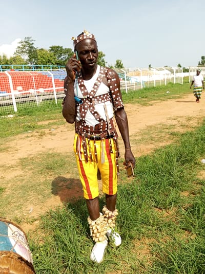 A man in traditional attire stands on a grassy field in Takau, Nigeria, engaged in a phone call. He wears a white t-shirt under a brown leather harness adorned with white buttons, yellow shorts with red stripes, and white shoes with leg coverings made of small, light-colored gourds or seeds. A matching headpiece is also visible. He holds a flip phone to his ear and another phone in his other hand. A drum rests on the ground to his left. In the background are metal bleachers and a person walking along a dirt path. The environment is predominantly green and brown with a clear sky above.