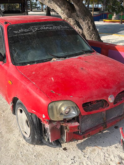 An old, red, damaged car sits outside on a concrete surface. The front of the car is heavily dented, with the radiator exposed and the headlight cracked. The windshield is dusty, marked with a sticker, and a tree branch overhangs the left side of the vehicle. Overall, the car appears to be in a state of disrepair.