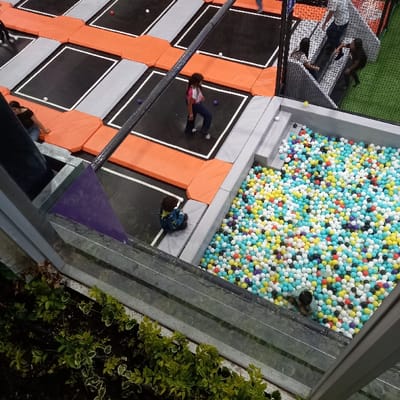 The image depicts an indoor recreational facility, specifically a trampoline park and ball pit, located in Tumbaco, Ecuador. The scene is viewed from an elevated perspective, looking down through a clear barrier, with green foliage visible below the barrier in the foreground.
The facility features numerous interconnected trampolines with black jumping surfaces outlined in white, separated by orange and grey padded safety mats. Several individual plastic balls, colored red, white, and purple, are scattered across the trampoline surfaces. In the middle ground, a female with long dark hair, wearing a white and pink top with blue pants, stands centrally on a trampoline. To her left, another individual in a dark top kneels on the orange padding between trampolines.
To the right, a large rectangular ball pit is filled with a dense collection of multi-colored plastic balls, including white, light blue, yellow, red, purple, black, and green. A person, partially submerged in the balls, is visible in the lower right portion of the ball pit. Adjacent to the ball pit, a small child in a dark blue patterned jacket sits on a grey padded edge.
In the background-right, a netted climbing structure with a green padded floor is visible. Several individuals are present in this area; one person in a white shirt and dark pants stands, another in a dark top moves, and a third is seated within the structure. The overall environment appears well-lit and designed for recreational activity.