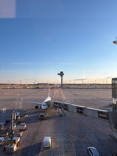 This is a photograph taken from an elevated perspective, likely a window at an airport, looking out onto an airport tarmac. The scene depicts a large passenger airplane connected to a jet bridge, with ground support equipment and other aircraft visible in the background. The sky is clear blue with a hint of sunset colors near the horizon, indicating late afternoon or early evening.

The main subject is a white airplane with red and blue accents on its tail. It is parked at a gate, with a silver and white jet bridge extending to its fuselage. The jet bridge has "Revolut" branding visible. In front of the plane, a white baggage tug is positioned.

To the left of the plane, several cargo containers and baggage carts are lined up on the tarmac. A signpost with green symbols and the text "B08" is visible amongst the cargo. Further left, more aircraft are parked in the distance.

In the middle ground, the distinctive control tower of Schönefeld Airport (now part of Berlin Brandenburg Airport) stands tall against the sky. Its skeletal structure is clearly visible. Various airport buildings and hangars are present in the background, along with a line of tall light poles.

On the right side of the image, a portion of a modern airport terminal building is visible, with large glass windows. A silver car is parked on the tarmac in the lower right corner of the frame. The tarmac itself is marked with yellow and red lines indicating taxiways and parking positions. The overall environment is that of a busy international airport, with a sense of calm due to the subdued lighting of the setting sun. There are no visible people actively engaged in interaction, though their presence is implied by the operational nature of the airport.