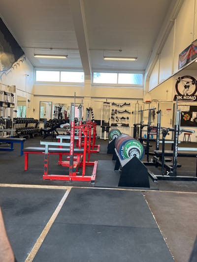 An interior wide-angle photograph depicts a well-lit, unoccupied weightlifting gym identified as "VK KONO" in Odense, Denmark. The facility features a light-colored ceiling with exposed support beams and multiple fluorescent strip lights. High, rectangular windows along the upper back wall allow natural light. The flooring consists of dark rubber mats with visible seams.

Along the left wall, a mural with abstract blue and grey wave-like patterns is present. Below it, multiple dumbbell racks with a range of free weights and various fitness machines are visible. In the midground, several power racks and squat racks are arranged on the rubber matting. The racks on the left are predominantly red-framed, while those on the right are black-framed; both types bear the vertical inscription "POWER EQUIPMENT." Associated with these racks are multiple flat benches with black padding and red or light-colored frames.

Further right in the midground, weight plate storage racks hold numerous "ROGUE" brand bumper plates in various colors (green, blue, red, yellow, white). Barbells are present, some stored vertically against walls or within racks. The far right wall displays a prominent circular banner featuring the "VK KONO" logo, a weightlifter graphic, and text that includes "Odense" and "Vægtløftning og Styrkeløft." Below this banner is a dark board or display and a small red stool. Various types of weightlifting bars and attachments hang on the back wall. No individuals are visible engaging in activities. A partial view of a limb is visible at the bottom left, suggesting the vantage point of the photographer.