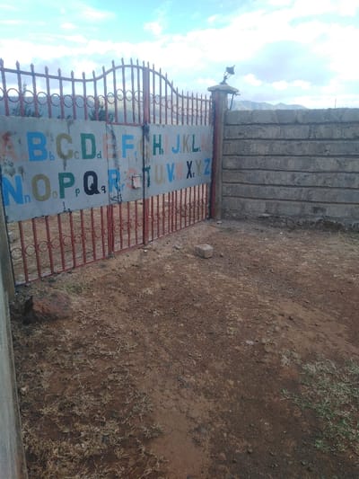 A red metal gate with an alphabet sign hangs between two pillars. The sign displays colorful letters from A to Z. Beyond the gate, a weathered gray brick wall stands. The ground is predominantly dirt with sparse vegetation. The sky is a muted blue with scattered clouds and mountains in the distance.