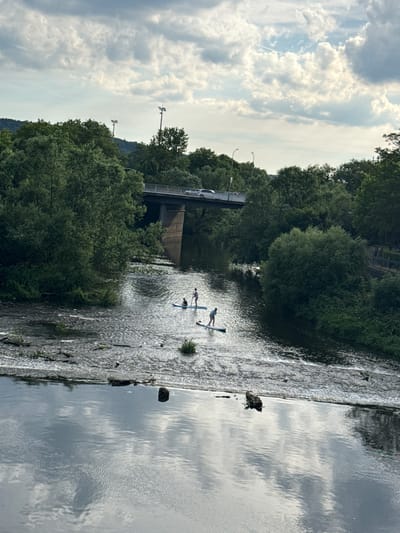 The image depicts a scenic river scene with people paddleboarding. In the foreground, several individuals are seen on paddleboards, navigating the calm waters. The river is bordered by lush green trees on both banks, creating a natural and tranquil setting.

A notable feature in the background is a bridge, likely part of a road, with cars visible on it. This suggests an urban or semi-urban environment where nature and infrastructure coexist. The sky is partly cloudy, with the sun filtering through, indicating it is likely daytime. The overall atmosphere appears peaceful and recreational, with the individuals enjoying a leisurely activity on the water. There are no visible text elements in the image. The location is Wetzlar, Germany.