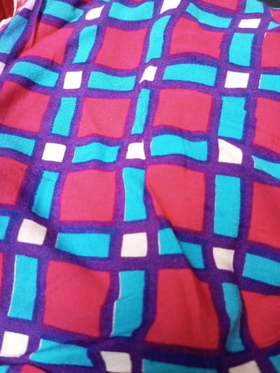 The image depicts a textile with a vibrant, abstract grid pattern. The grid consists of purple lines that form uneven squares. Inside the squares, there are alternating blocks of red and teal. Small white squares are interspersed randomly at the intersections of the purple gridlines. The fabric has folds and wrinkles, giving the surface a textured appearance. The colors are rich and saturated.