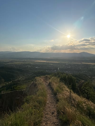 The image depicts a panoramic landscape view captured from a high elevation in the vicinity of Stob, Bulgaria. The time of day is characterized by a low-angle sun in the upper right quadrant, producing distinct lens flares and sunbeams. The sky transitions from light blue overhead to a warmer, lighter hue near the horizon, featuring scattered white and grey clouds, some partially illuminated by the sun.
In the immediate foreground, a narrow, unpaved dirt path, approximately 1-1.5 meters wide, traverses a ridge. The path is flanked by tall, dry, golden-brown grasses and some greener foliage, and it curves subtly towards the right of the frame. To the left of the path, a steep, eroded slope descends sharply. A section of a light-colored wooden fence or railing is partially visible on a lower ledge on the left, indicating a potential overlook or managed area. No human figures, animals, or specific objects are discernible on the path or the immediate foreground.
The midground displays an expansive valley system. Below the foreground ridge, densely forested slopes transition into a broad, relatively flat agricultural plain. This plain is composed of a mosaic of cultivated fields, appearing in varying shades of green and yellowish-brown. A narrow, serpentine body of water, likely a river or stream, is visible cutting across the plain from left to right.
The background features a range of hazy, undulating mountains forming the distant horizon. Below these mountains, a settlement, consistent with the description of Stob village, is visible within the valley. It comprises clusters of buildings and what appear to be roads, surrounded by a dense canopy of trees. The overall atmosphere indicates clear weather with some natural atmospheric haze.