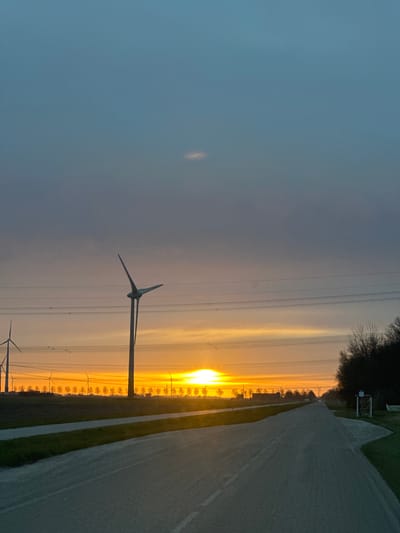 The image depicts a landscape with a road on the right, a field in the middle, and a cloudy sky on the left, transitioning to a bright sunset on the horizon. A row of trees is visible along the field. Electrical pylons with wires run across the scene. Two wind turbines are visible, one in the foreground and one in the background. The location is likely in the vicinity of Lelystad, Netherlands.