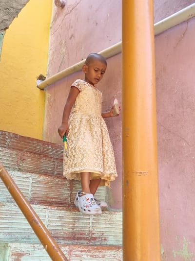 A young child with a shaved head and a red bindi on her forehead stands on a set of weathered stairs in Pedda Kammavaripalli, India. She is dressed in a light-colored, patterned lace dress with short sleeves and white Crocs-style sandals adorned with charms, and a silver anklet on her left ankle. The child holds a yellow and green screwdriver-like tool in her right hand and a small, clear plastic bottle containing a granular substance in her left hand. She is looking downwards and slightly to her left. The immediate environment includes a faded pink wall to her right and behind her, displaying cracks and texture. A bright yellow wall is visible to her left. A horizontal light-colored pipe runs along the pink wall near the top. A prominent vertical orange-brown pipe or pillar is positioned in the foreground on the right side of the image. A diagonal orange-brown pipe, likely a handrail, is visible in the lower left foreground. The stairs feature peeling light blue-green paint over a reddish-brown, textured brick or tile pattern.