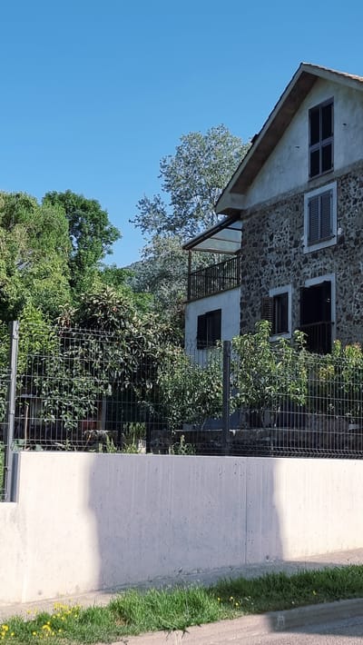 The image depicts a close-up view of a building with stone walls and dark-shuttered windows, set against a clear blue sky. The building appears to be a house or part of a larger structure, with a slanted roofline and an exterior balcony with metal railings. Lush green foliage, including trees and bushes, surrounds the building, with some of the greenery visible behind a metal fence. To the right of the building, a concrete path runs alongside a strip of vibrant green grass dotted with small yellow wildflowers. The overall scene suggests an outdoor setting, possibly a garden or courtyard, in what appears to be a rural or semi-rural location. The lighting suggests it is daytime, with shadows cast on the concrete path indicating the sun is relatively high. No people are visible in the frame. There is no visible text in the image. The location context provided is Sant Joan les Fonts, Spain.