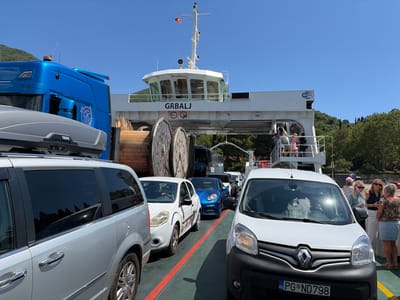 This is a photograph taken at the Lepetane ferry terminal in Montenegro. A ferry named "GRBALJ" is docked, carrying several vehicles and passengers. The ferry is white with a distinctive bridge structure at the front. A large blue truck is parked on the ferry, partially obscuring a grey minivan. Other cars, including a white sedan and a blue sports car, are also lined up. A white van is in the foreground. Several people are visible on the ferry deck and on a raised walkway, some interacting with each other. The environment appears to be a sunny day with a clear blue sky. The background shows green hills and trees, indicating a coastal or mountainous region. The presence of large wooden spools on the ferry suggests it might also transport cargo.