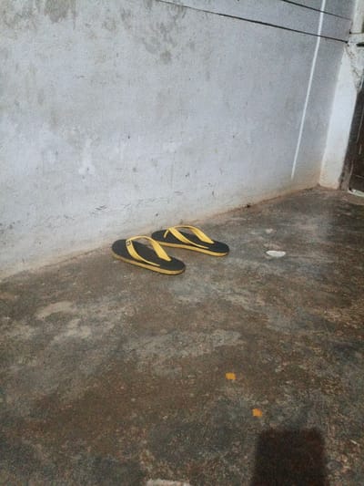 A pair of black and yellow flip-flops are placed on a dirty, wet concrete floor in front of a rough, gray concrete wall. The scene appears to be outdoors, possibly in a low-income area or alleyway within Meunasahuket, Indonesia. There are a few small pieces of debris scattered on the floor, including a white crumpled piece of paper and two small yellow squares. The lighting is dim, and the overall impression is one of neglect.