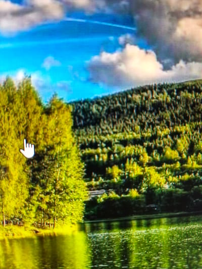 The image shows a picturesque landscape, likely in the city of Vistino, Russia, with a focus on the natural environment. In the foreground, a body of water reflects the vibrant green and yellow hues of the surrounding trees and the bright blue sky. The trees on the left side of the image are a prominent feature, with their foliage appearing in shades of bright yellow and green. A white cursor in the shape of a hand is visible on the left side of the trees. Behind the trees, a densely forested hillside stretches across the middle ground, with a mix of dark green and lighter green tones, suggesting a variety of trees and possibly varying levels of sunlight. The sky above is a brilliant blue, punctuated by fluffy white and gray clouds, with hints of yellow and pink, suggesting it may be late afternoon or early morning. The overall scene conveys a sense of natural beauty and tranquility.