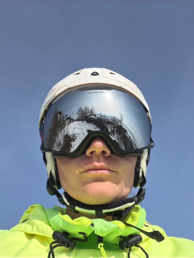 A close-up portrait depicts an individual wearing ski equipment against a clear blue sky. The person wears a white Atomic-branded ski helmet and silver-mirrored Blizzard ski goggles, which reflect a panoramic view of a snow-covered mountain landscape. The reflection includes bare trees, exposed rocky terrain, a bright sun glare, and a distant ski lift structure. The lower section of the individual's face, encompassing the nose, mouth, and chin, is visible beneath the goggles. They are dressed in a neon yellow-green ski jacket featuring black drawstrings and toggles around the collar. The scene depicts a high-altitude outdoor environment consistent with a ski resort in the city of Zelenaya Polyana, Russia.
