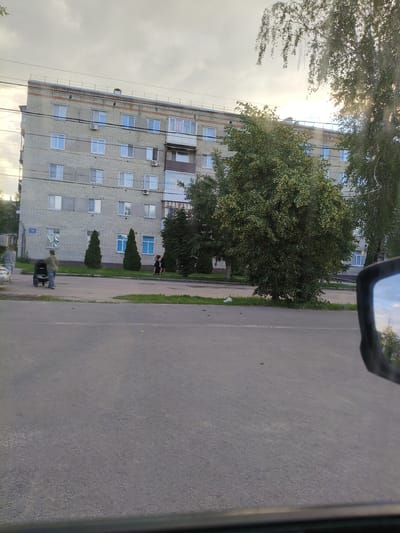 The image captures an outdoor scene in Petrovsk, Russia, with a multi-story apartment building as the central subject. The building appears to be constructed of light-colored bricks and has numerous windows. A balcony is visible on one of the upper floors, with a sign that reads "Продаю" (for sale) and "Квартиру" (apartment).
In front of the building, there is a paved area with a sidewalk and some green grass. Several trees are present, including a large, bushy tree to the right of the building and two conical evergreen trees near the entrance.
There are a few people in the image. Two women are walking on the sidewalk. One is pushing a stroller, and another is walking behind her. Further to the right, two figures are visible near the building, though their activities are unclear due to their distance and the quality of the image.
The scene appears to be on a street, with a paved road in the foreground. A car is partially visible on the left side of the road. The sky is overcast with some hints of sunlight breaking through, suggesting it might be late afternoon or early evening. The overall mood of the image is everyday and observational, with no specific event being highlighted. The reflection in the car's side mirror is also visible, showing a portion of the sky and trees.