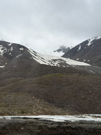 The image shows a rocky, mountainous landscape in Kyrgyzstan under a cloudy sky. In the foreground, there is a band of melting snow and ice with some dark, wet earth. Behind this, a steep, rocky slope rises, sparsely covered with patches of green vegetation. Further up, the mountain is covered with scree and patches of snow. In the background, a large glacier is visible, partially obscured by clouds. The overall impression is one of a remote, harsh, and natural environment. There are no people or man-made structures visible in the image.