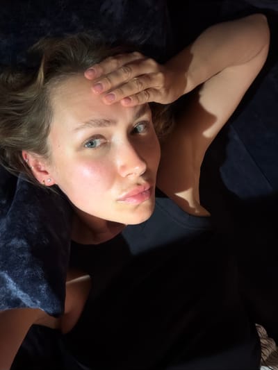 A woman with light brown hair lies on a dark surface, possibly a couch or bed, with her left arm bent and resting on her forehead. Her right arm is bent and positioned behind her head. She is looking upwards towards the camera with a neutral expression. The lighting is dim, with a spotlight creating strong shadows. She is wearing a dark top. The scene appears to be indoors, with no discernible outdoor or city elements visible. No specific location in Mühlbach - Rio di Pusteria, Italy can be identified from the image.