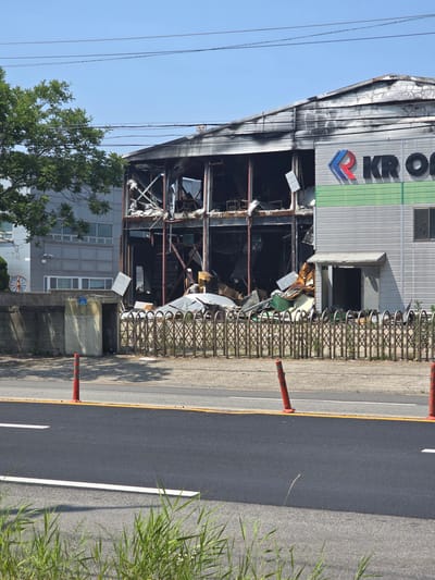 The image shows the exterior of a severely damaged building in Pyeongtaek-si, South Korea. The building's upper floors are extensively burned and collapsed, with exposed metal framing and debris visible. The lower section of the building appears to be intact but shows signs of smoke damage. A large sign with red and blue lettering, reading "KR O" (likely part of a company name), is visible on the undamaged portion of the building. A metal fence with a diamond pattern stands in front of the building, separating it from a road. Orange traffic cones are placed on the roadside. The sky is clear and blue, and a tree with green foliage is to the left of the building. The overall scene suggests a recent fire or explosion.