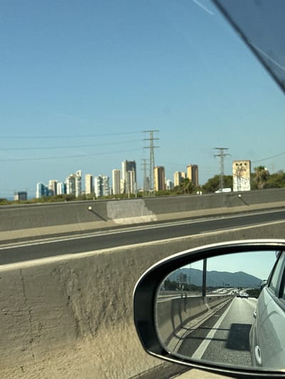 The image captures a view from a vehicle, likely on a highway, with a city skyline visible in the distance. The foreground shows a concrete barrier alongside a multi-lane road, reflecting the clear blue sky. In the middle ground, a collection of modern high-rise buildings, characteristic of a city, are spread across the horizon. These buildings vary in height and architectural style, suggesting a developed urban area. Several tall power pylons are also present, indicating the presence of significant electrical infrastructure. To the right, a somewhat weathered, rectangular structure with graffiti is visible. Palm trees are scattered amongst the buildings and pylons. The far distance features a range of mountains under a bright sky. The reflection in the car's side mirror offers a glimpse of the road ahead, showing other vehicles and the same mountainous backdrop, confirming the mountainous coastal setting. This scene is consistent with the appearance of Benidorm, Spain, a city known for its distinctive high-rise architecture and proximity to mountains. No people are directly visible in the image.