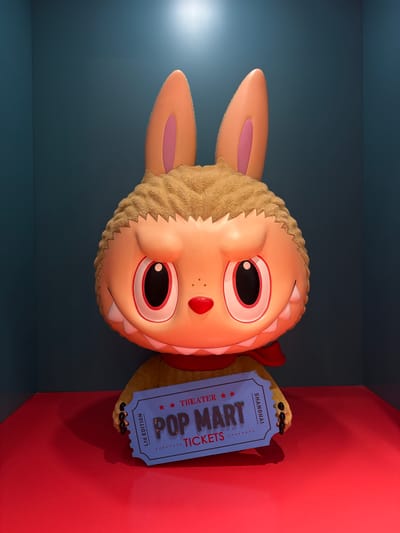 A plush toy with large ears, a fuzzy brown head, and an orange face with large, wide eyes and sharp teeth is displayed against a dark blue background. The toy is wearing a red scarf and holding a blue ticket that reads "POP MART THEATER TICKETS SHANGHAI LTD EDITION." The ticket is positioned in front of the toy's lower body. The scene appears to be an indoor display, possibly in a store or exhibition space, with a red platform beneath the toy. The lighting suggests an indoor setting with a spotlight from above. No people are visible in the image. This display is located in Pudong, China.