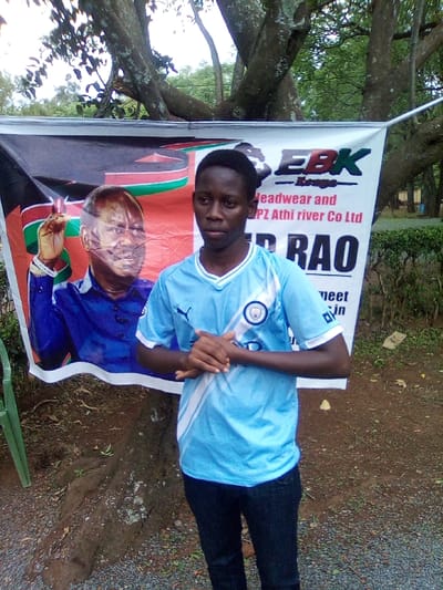 In South Sakwa ward, Kenya, an outdoor scene depicts a young male standing in front of a promotional banner. The male, with dark skin and short dark hair, wears a light blue football jersey with white accents and dark trousers. His hands are clasped together at waist level, and he looks directly forward. Behind him, a large banner is suspended, partially obscured by his body. The left side of the banner displays a prominent image of a man in a blue shirt, with the Kenyan flag colors (black, red, green, white) forming a background, and his right hand raised. The right side of the banner features a white background with black text, including "EBK" with a logo, "Headwear and PZ Athi river Co Ltd," and "RAO." A large tree trunk with visible roots is present behind the banner and to the male's left. The ground consists of dirt and gravel. A green plastic chair is partially visible on the far left. The overhead environment shows tree branches and an overcast sky.
