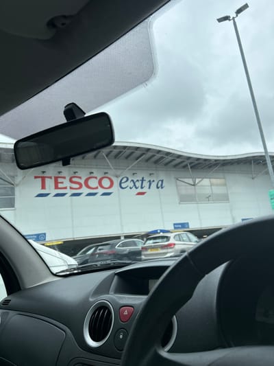 The image, captured from the interior of a vehicle, displays the exterior of a Tesco Extra supermarket and its associated car park in Havant, United Kingdom. The lower portion of the frame shows the vehicle's dashboard, including a black steering wheel on the right, an air vent with a metallic ring, and a red hazard light button. A rearview mirror is suspended from the windshield, which features a stippled pattern in its upper section.

Outside the vehicle, a car park occupies the midground, containing several parked automobiles, including a dark-colored estate car and a silver hatchback. A blue "Parking" sign with a directional arrow is partially visible above the parked vehicles. The background is dominated by the large, white facade of the Tesco Extra building, featuring its distinctive "TESCO extra" logo in red and blue lettering. The building exhibits an undulating roofline and rectangular windows. A tall, grey lamppost with two luminaires stands on the far right against a uniformly overcast, grey sky. No persons are visible within the frame.