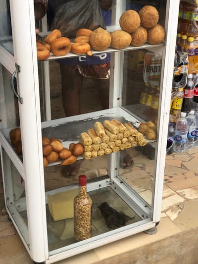 The image depicts a mobile glass display cabinet positioned on a paved surface in Egan, Nigeria. The cabinet, constructed with a white frame and transparent glass panels, features three shelves filled with food items.
On the top shelf, approximately eight golden-brown ring-shaped doughnuts are arranged on the left, while five spherical, golden-brown fried dough balls are stacked on the right. Below this, the middle shelf displays a pile of irregular, golden-brown fried items, possibly fritters, on the left. To their right, a pyramid stack of approximately 30-40 light golden-brown rolled items, resembling spring rolls, occupies a significant portion of the shelf. A smaller cluster of additional fried items is visible further right. The bottom shelf contains a large, pale yellow rectangular block on the left, a clear glass bottle with a red cap filled with groundnuts (peanuts) in the center, and a dark, crumpled object, possibly a plastic bag, on the right.
Reflections within the cabinet glass show the lower torso and legs of at least two individuals. One person, wearing dark shorts with a patterned hem, is visible behind the top and middle shelves, with their feet appearing on the paved ground. Another set of feet, clad in sandals, is reflected near the bottom shelf. A hand is partially visible in the top left corner, seemingly interacting with items inside the cabinet.
To the right of the display cabinet, an adjacent stall or display area is visible, featuring various bottled beverages including water, Coca-Cola, Fanta, and Maltina, some packaged in clear plastic bags. The ground beneath the cabinet and extending to the right is composed of irregularly shaped tan and brown paving stones.