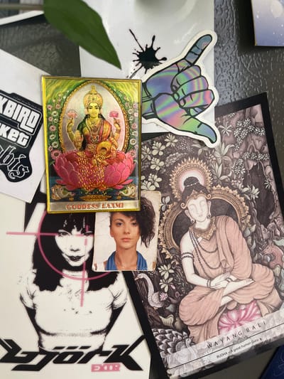 A high-angle, close-up shot showcases a collection of items on a dark, reflective surface, possibly a table. The arrangement includes several stickers and a small framed image.
Dominating the center of the frame is a sticker of a hand forming the "shaka" sign, rendered in a holographic, iridescent material that shifts through shades of purple, blue, green, and yellow. To its left, a black ink splatter sticker is visible.
To the right of the shaka sticker, a small, rectangular framed image depicts a Hindu deity, likely Lakshmi, adorned with jewelry and holding lotus flowers. The image has a golden frame with intricate floral details. The text "GODDESS LAXMI" is written vertically along the right edge of this frame in a red font.
Overlapping the framed image and extending upwards is a larger print or poster featuring a detailed, intricate illustration in muted tones. The artwork depicts a figure lying down, surrounded by decorative motifs and what appear to be floral elements and possibly animals. Text is visible along the right edge of this print: "BUDDHA - A great human Guru. WAYANG GULI BALI".
Below the framed image and partially obscured by it is a small photograph of a young person with dark hair, looking towards the viewer. Their attire includes a blue garment.
Further down and to the left, a sticker with bold, outlined text in black and white is partially visible, with the letters "BIRD" and "KER" discernible.
The overall scene is an eclectic arrangement of pop culture and spiritual imagery. The lighting suggests an indoor setting, and no specific time of day or weather conditions are evident. The location context provided is Балаклавский округ, Ukraine, but there are no visual cues in the image that confirm this.