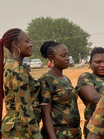 Three young women wear camouflage clothing outdoors. The woman on the bottom left has a large gold hoop earring and red braids. The woman above her has black braids and a teardrop-shaped gold earring. The woman on the top right has dreadlocks and appears to be standing behind the other two. Several cars and a tree are visible in the background.