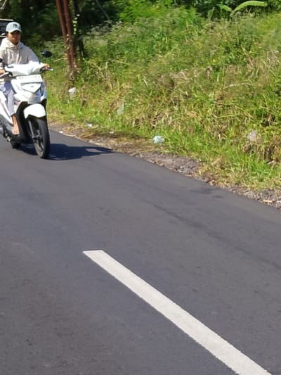 A young man wearing a light-colored hoodie and a baseball cap is riding a white scooter on a paved road. He is facing to the left, and his body is partially obscured by the scooter. The scooter has a license plate that appears to read "DK 4773 FU." The road is asphalt and has a white line painted on it, dividing the lanes. To the right of the road is a dense area of green vegetation, likely grass and bushes. In the background, there are a few vertical brown poles. The scene is illuminated by bright sunlight, casting shadows on the road. The image was taken in the city of Tabanan, Indonesia.