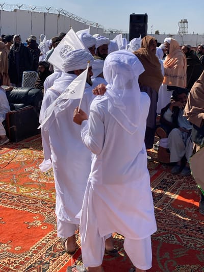 The image depicts an outdoor gathering in Mandisar, Afghanistan, under bright daylight. A crowd of predominantly men is present, many dressed in white shalwar kameez and turbans or other head coverings. Several individuals are holding small white flags mounted on sticks, which display a black circular emblem containing script and a central design, reminiscent of the Shahada and the Seal of Prophet Muhammad. The ground is covered by large, ornate red and black patterned rugs.
In the foreground, two men in white shalwar kameez stand facing each other; one holds a flag in his right hand. To their left, part of a black leather sofa or similar seating is visible. In the background, a large speaker is mounted on a stand. Beyond the crowd, a grey wall topped with barbed wire fencing is visible under a clear blue sky. Further in the background, a metal lattice tower is present. Individuals in various traditional garments, including some with brown shawls or blankets, are scattered throughout the scene. One man in the mid-ground is seated on the ground, leaning against what appears to be a stringed instrument. The overall scene suggests a public assembly or celebration.
