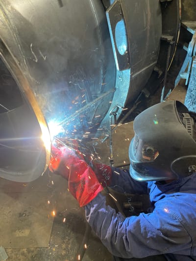 A welder in Zaljevo, Montenegro, is welding a large metal object. The welder is wearing a dark-colored welding helmet, a blue long-sleeved shirt or jacket, and red gloves. Sparks are flying from the welding arc. The metal object appears to be curved or rounded. The floor is visible in the lower right of the image, showing signs of being a workshop floor.