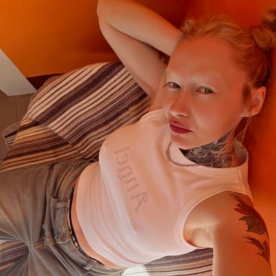 A young woman with blonde hair styled in a bun is lying on a striped blanket over what appears to be a vibrant orange surface. She is wearing a white tank top with the word "Angel" spelled out in rhinestones and a pair of faded denim jeans. Tattoos are visible on her neck, upper arm, and forearm. She is wearing a necklace that says "BABY". Her head rests on her left hand, and she is looking up at the camera. The setting may be indoors. The location provided, Тэльмінскі сельскі Савет, Belarus, is not directly evident from the image itself.