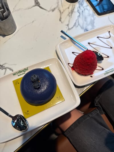 The image displays an overhead shot of two colorful desserts on a white marble table, likely inside a cafe or dessert shop in Hanau, Germany.

In the upper portion of the frame, a white rectangular plate holds a vibrant red, dome-shaped dessert, possibly a sorbet or mousse, characterized by a textured, velvety surface. It is garnished with a zigzag drizzle of dark chocolate sauce. A silver spoon rests on the plate next to it. Green text, "Seydioğlu BAKLAVA," is visible along the edge of this plate, indicating the establishment.

Below this, a second white rectangular plate features a striking blue, dome-shaped dessert, possibly a blueberry mousse cake, topped with two fresh blueberries. This dessert is presented on a square yellow paper, which also prominently displays the green text "Seydioğlu BAKLAVA" in a rotated orientation, along with a smaller "Seydioğlu BAKLAVA" logo. Another silver spoon is placed on this plate.

To the left, a black smartphone is partially visible on the table. In the lower-left corner, a cylindrical metal container holds various paper items, including sugar packets, with "Zucker" (German for sugar) partially legible on one, and "UCC Trading Dubai" on another.

In the blurred background, the legs and lap of a person wearing dark grey pants or jeans are visible, indicating a patron seated at the table, enjoying the desserts. The scene suggests a pleasant outing, focusing on the visually appealing sweet treats.