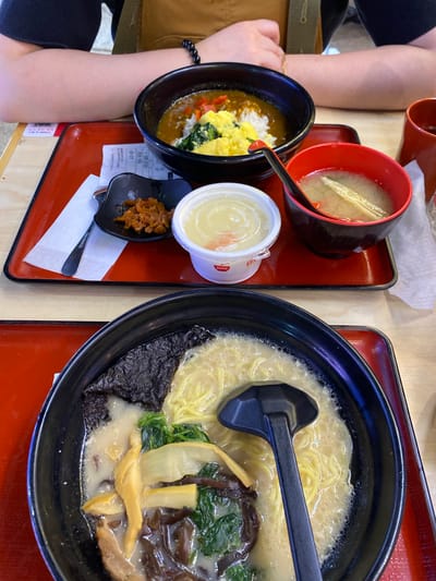 This is a close-up, eye-level shot of two Japanese meals on red trays on a wooden table. The meals are placed side-by-side. The tray in the background contains a bowl of curry rice with scrambled eggs and spinach, a small dish of pickled vegetables, and a bowl of miso soup. A person's arms are visible in the upper part of the frame. The tray in the foreground contains a bowl of creamy broth ramen with noodles, seaweed, mushrooms, and green vegetables. A black ladle rests in the ramen. This scene is at a restaurant located in Qingyang District, China.