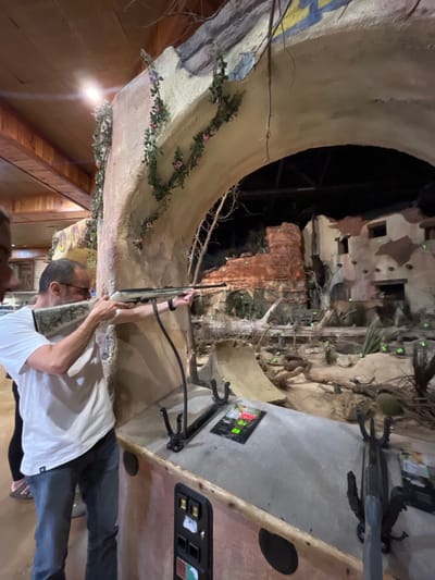 The image depicts a man indoors, holding a rifle in what appears to be a shooting gallery or simulation. He is wearing a white t-shirt and jeans. The background includes a display of a simulated environment with rock formations, buildings, and trees. The scene is designed to resemble a natural or historical setting. The presence of a digital display panel and gun mount suggests this is a simulated shooting experience. The location is possibly within a retail establishment in the city of San Antonio, United States of America, known for attractions and immersive experiences.