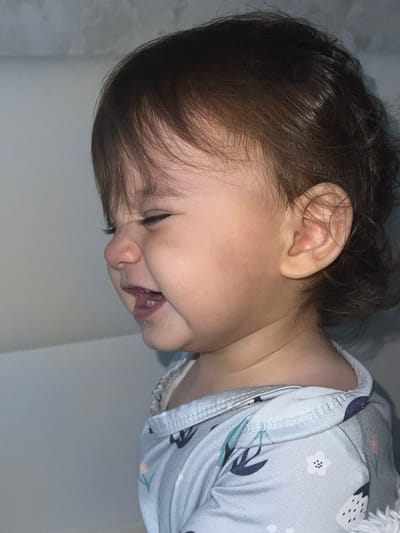 Forensic analysis of the image, captured in Elena, Bulgaria, presents a close-up profile view of an infant or toddler. The subject's eyes are tightly squinted shut, and the mouth is open, revealing lower deciduous teeth and the tongue. Dark brown hair frames the face and partially covers the left ear. The child is wearing a light-colored, long-sleeved garment adorned with small floral and leaf patterns. The immediate background is an indistinct, light grey wall or surface, with a hint of a white, soft-textured object at the bottom right.