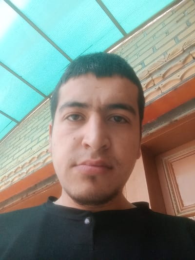 The image depicts a close-up portrait of a male subject, located in Kasan, Uzbekistan. The individual has short, dark hair, brown eyes, and stubble on his chin and upper lip. He is wearing a black collared shirt with a visible silver button on the placket, looking directly at the camera. The background consists of a translucent light-blue corrugated roof structure with metal beams above the subject. To the upper right, a light-colored brick wall with decorative relief patterns is visible. A section of an orange-brown wooden frame, possibly from a door or window, is present in the lower right background.