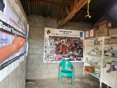 The image displays the interior of a simple building, likely a health center or aid station, in Nzulo, Democratic Republic of the Congo. The walls are constructed from concrete blocks, and the ceiling features a corrugated metal roof supported by wooden beams.

On the left, a person's hand points to a white banner partially visible, which clearly displays the text "Daniel Torsiello Health Center" and mentions "Health," along with an email "globalbrightenignalan." A larger central banner dominates the back wall, introducing "GLOBAL BRIGHT CHILDREN 'GBC' Ashi." This banner details "THE IMPLEMENTATION OF HUMANITARIAN ACTIONS TO ASSIST VULNERABLE PEOPLE IN THE DEMOCRATIC REPUBLIC OF CONGO, NORTH KIVU PROVINCE." It prominently welcomes "our Father Daniel Torsiello and Jackie Jaramilla," featuring their photograph alongside a collage of numerous smaller photos depicting various community activities. The year "2021" is also visible on this banner.

To the right, white shelving units are stocked with an assortment of medical supplies, including boxes, bottles, and packages, indicative of a dispensary or storage area for pharmaceuticals. Cardboard boxes are also stacked on top of and beside the shelves. A single green plastic chair sits on the concrete floor in front of the central banner. A yellow, string-like decoration hangs from a beam. The overall scene suggests a functional facility dedicated to humanitarian and health services.