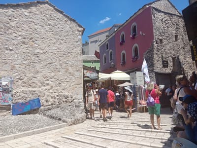 This media file captures a bustling scene in the historic old town of Mostar, Bosnia and Herzegovina. The image is dominated by the distinctive stone architecture of the region, with a prominent rough-hewn stone building on the left and colorful, multi-story buildings on the right, featuring pink and purple facades with arched windows adorned with flowers. A bright blue sky with scattered white clouds suggests a sunny day, likely mid-morning or early afternoon, given the strong shadows.
Several people are visible, appearing to be tourists, navigating a stone-paved walkway that slopes upwards. They are dressed in casual summer clothing, indicative of warm weather. Some are walking away from the viewer, while others are facing towards the camera. In the background, market stalls with awnings are set up, displaying goods and adding to the lively atmosphere. Signs in the image include "MUSEUM," "SAJAM ANTIKVITETA" (Antique Fair), and "Olimpija," suggesting commercial activity and cultural heritage sites. Another sign reads "THE OLD BRIDGE MUSEUM," a key landmark in Mostar. A blue board with graffiti is leaning against a stone wall on the left, adding a touch of urban art to the ancient setting. The overall impression is one of a vibrant tourist destination, showcasing the blend of historical architecture, local culture, and contemporary life on a beautiful sunny day.