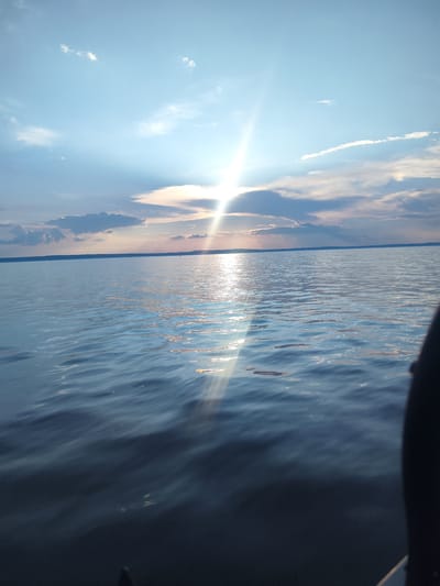 The image captures a serene view of a body of water, likely a lake or large river, under a bright, partially cloudy sky. The sun is positioned high in the sky, its rays creating a brilliant lens flare that extends across the scene. The water is calm with gentle ripples, reflecting the soft hues of the sky and the sunlight. In the distance, a dark, horizontal line indicates a distant shoreline, possibly with a silhouette of trees or land. The sky is a gradient of blue, transitioning from a lighter shade near the horizon to a deeper blue overhead, with scattered white and gray clouds. The overall impression is one of tranquility and natural beauty during daylight hours. There are no people, animals, or man-made structures clearly visible in the foreground or midground. The background suggests a natural landscape, consistent with an outdoor location such as the outskirts of Галево, Russia, where such bodies of water are common.